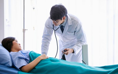 asian young male doctor wearing eyeglasses and white gown, using stethoscope, checking up health of sick female patient lying down on bed with blanket in ward recovery room in hospital