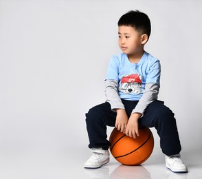 portrait of a preschool little smiling asian boy wearing a trendy raglan with a cool wolf print, sitting on a big orange basketball ball and waiting to be played with