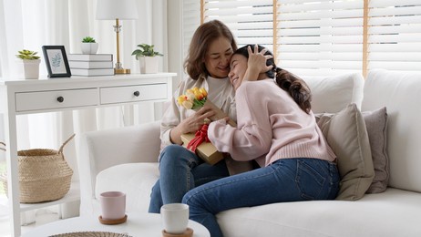 attractive beautiful asian middle age mum sit with grown up daughter give gift box and flower in family moment celebrate mother day. overjoy bonding cheerful kid embrace relationship with retired mom.
