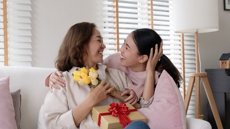 attractive beautiful asian middle age mum sit with grown up daughter give gift box and flower in family moment celebrate mother day. overjoy bonding cheerful kid embrace relationship with retired mom.