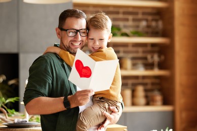 portrait of happy family father with excited child son hugging smiling  while celebrating fathers day together at home. little boy congratulating giving dad homemade greeting postcard