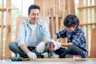 asian father look carefully son use drill in their workplace of carpentering with happy emotion. asian family concept to stay at home and enjoy good relationship hobby together.