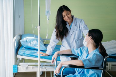 smiling young asian female doctor doing routine checkup and talking and comforting depressed patient sitting on wheelchair in hospital ward