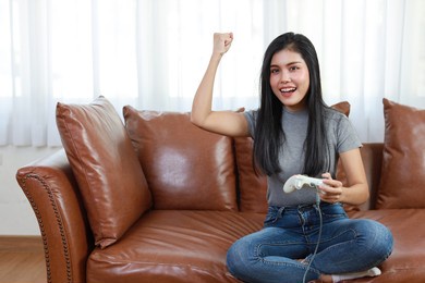 vdo game console station concept. active asian woman sitting on sofa, holding joystick and playing exciting game. cute girl looked excited with game controller console