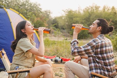 enjoy asian young woman, girl and man cheering with beer bottle, sitting on chair . adventure couple, people camping in forest. eco activity, lifestyle nature on holiday concept.