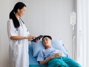 in hospital private room, one female doctor with stethoscope and laptop in her hands talking with patient man as he laying on bed with iv tube. look each other with smile. seems guy recovery soon