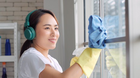 happy asian woman in headphones listening to music and cleaning window with cleanser at home, housework and housekeeping concept