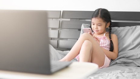 asian child learn or kid girl happy smile to shopping play by smartphone or study online and work from home on computer laptop by sit cross legged leaning pillow on bed for back to school e-learning