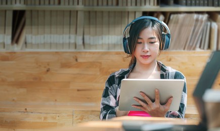 young beautiful asian woman with headphone using tablet for online learning while sitting in library.