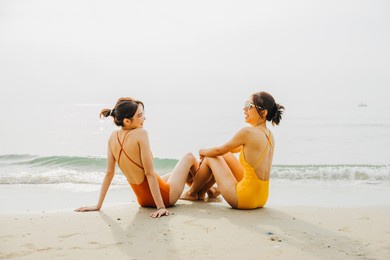 two beautiful asian women wearing orange and yellow bathing suits. they  were talking each other while sitting on the beach. summer and friendship concept.