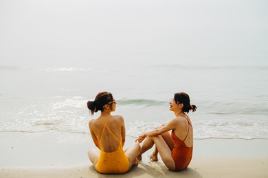 two beautiful asian women wearing orange and yellow bathing suits. they  were talking each other while sitting on the beach. summer and friendship concept.