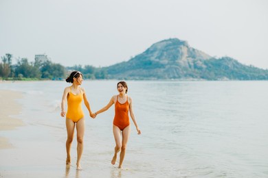 two beautiful asian women wearing orange and yellow bathing suits. they are running together on the beach. summer and friendship concept.