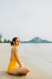 a cute asian woman in an yellow swimsuit is sitting on a beach. she is relaxing on summer vacation.