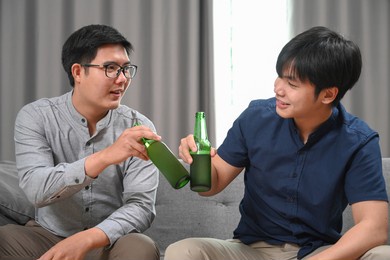two asian man clinking their glasses of beer while sitting on sofa at home.