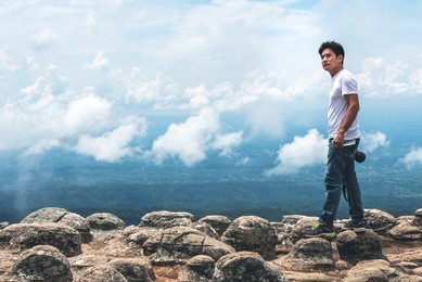 asian photographer middle-aged man standing on a rocky mountain which looks like a button, he is looking up, with view of clouds on blue sky background, to people and travel on vacation  concept