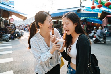 two asian chinese friends drinking cold beverage by straw in disposable plastic cup while standing on road in local traditional market in thailand bangkok. smiling girls sisters sharing together