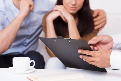 sign here. close-up of thoughtful young couple sitting on the couch while man in formalwear pointing clipboard with document 