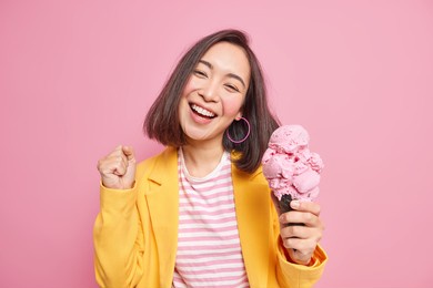 joyful adorable teenage girl with eastern appearance dark hair tilts head clenches fist eats tasty ice cream smiles broadly has fun enjoys summer dressed in stylish clothes isolated on pink wall
