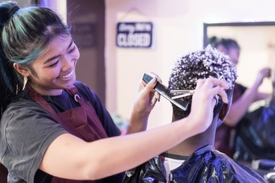 young asian woman barber  cuting black an white hair customer. beautiful hairstylist trimming scissors in hair cut salon shop.