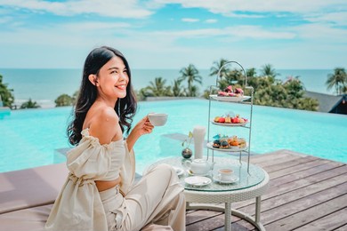 beautiful portrait young asian woman with afternoon tea set with coffee sit on chair around swimming pool in hotel resort