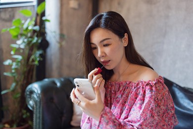 beautiful asian woman applying makeup cosmetics while looking at camera of mobile phone in modern cafe or home