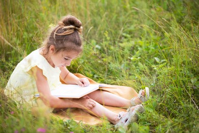 girl in a yellow dress sits in the grass on a blanket in a field and reads a paper book. international children's day. summer time, childhood, education and entertainment, cottage core. copy space