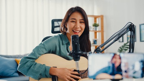 teenage asia girl influencer play guitar music use microphone record with smartphone for online audience listen at home. female podcaster make audio podcast from her home studio, stay at home concept.