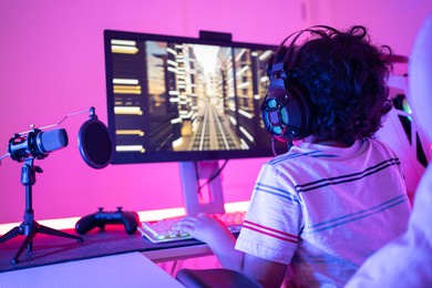 back of cute curly-haired little asian boy gamer wearing headphones sitting playing video games on a personal computer with rgb keyboard next to joystick and desktop microphone in a violet neon room.