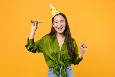 funny cheerful beautiful young brunette asian woman wearing basic green shirt birthday hat hold pipe dancing clenching fist looking camera isolated on bright yellow colour background, studio portrait
