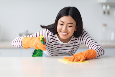 smiling asian lady cleaning kitchen table with spray and dust cloth, happy with her job, closeup, copy space. pretty young chinese woman house-keeping after moving to new apartment