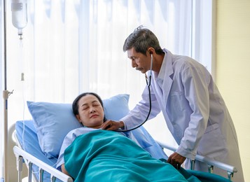 old senior elderly asian gray hair and mustache doctor in white lab coat using stethoscope monitor listen heartbeat pulse of female patient who sleep on hospital bed with normal saline solution drip.
