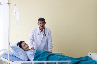 asian male elderly gray hair and mustache doctor in white lab coat uniform with stethoscope visit female patient lay down smile on bed cover by blanket inject with normal saline solution drip in ward.