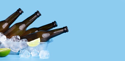 refreshing drink for summer party. full dark bottles of beer, without labels with flowing drops, lie in tray in ice cubes and pieces of lime, isolated on blue background, panorama, studio shot