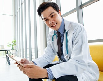 side view of busy asian male practitioner sitting in medical room in hospital and browsing mobile phone and laugh with happy and fun in relax time.