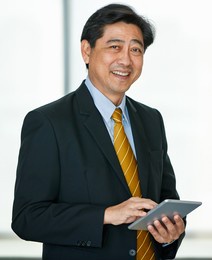 portrait 50s asian male executive visionary business male owner wearing formal suit and necktie, standing indoor office, using technology tablet, touching the screen, smiling confidently and reliably