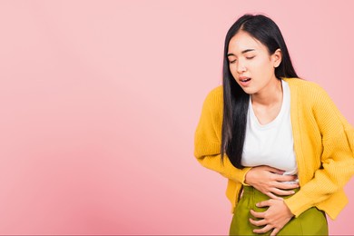 portrait of asian beautiful young woman has stomachache, female abdominal pain suffering from stomach ache, studio shot isolated on pink background, health and medical gastritis concept