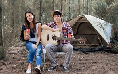adorable sweet asian adult two people or couple sitting at tent in forest for camping, smiling with happiness and freedom, playing guitar and singing in summer vacation while looking at camera