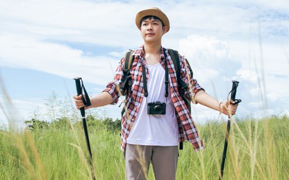 handsome asian young male traveler with camera, backpack, casual check shirt holding stick for trekking and hiking the mountains, standing in green field with happiness, freedom in summer vacation