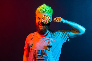 happy positive guy in stylish overalls and t shirt, posing with candy, covering his eye with tasty lolly pop, expressing optimism, having fun. colorful neon light, indoor studio shot.