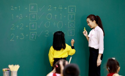 portrait shot of asian beautiful female mathematic teacher standing smiling cheer up while little smart schoolgirl student solving math equations on chalkboard in front of classmates in classroom.