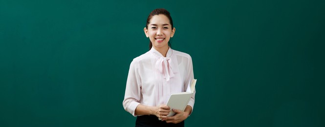 portrait shot of asian beautiful female teacher lecturer standing holding book in hands look at camera in front of chalkboard teaching little elementary school boy and girl children sitting on chairs.