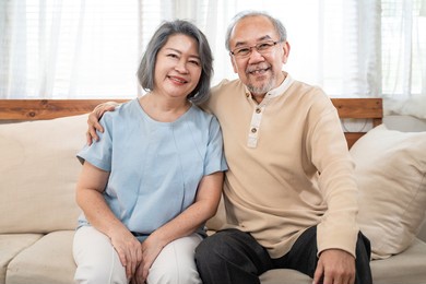 portrait of asian happy family, senior elderly couple enjoy retirement life at house. older grandfather and grandmother sit on sofa and smiling, looking at camera at home. health care activity concept