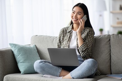 pretty young asian woman having phone conversation, sitting on couch in living room and using laptop, looking at copy space. cheerful lady resting at home, websurfing and talking on mobile phone