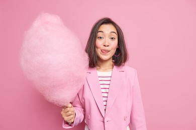 good looking brunette asian woman licks lips as holds appetizing sweet candy floss dressed in fashionable formal outfit isolated over pink background. teenage girl with delicious cotton candy