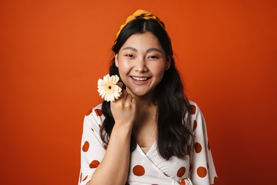 smiling young asian woman in summer clothes standing over orange background, holding flower