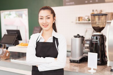 left side angle of asian barista woman crossed arms and happy standing to welcome customers with copy space in cafe coffee shop. barista work at coffee bar and food service business