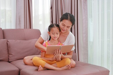 asian mother and daughter reading a book on the sofa. happy loving family at home