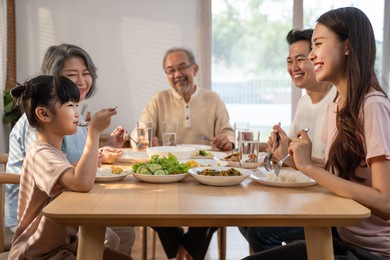 big asian happy family spend time having lunch on dinner table together. little kid daughter enjoy eating food with father, mother and grandparents. multi-generation relationship and activity in house