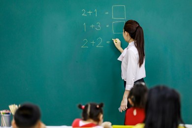portrait shot of asian beautiful female mathematic teacher standing holding chalk writing elementary school math equation questions on chalkboard while group of students sitting in blurred foreground.