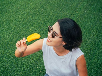 woman with popsicle. happy beautiful asian woman short hair wearing sunglasses and casual white sleeveless shirt holding yellow popsicle on green grass background, summertimes.
